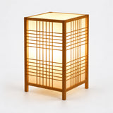 Luminária de mesa Shoji em bambu acesa, com trama geométrica e fundo cinza claro.